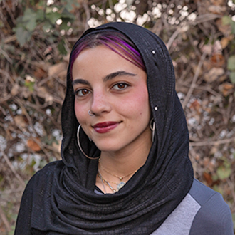 Nadin wearing a black headscarf and silver hoop earrings with trees in the background.