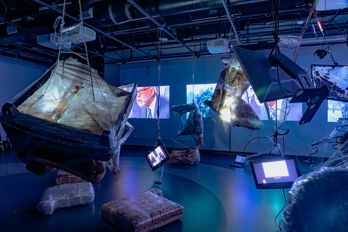 A multi-media room with a combination of images projected on the walls, active monitors hanging from the ceiling, and large objects wrapped in plastic placed strategically about the room. 