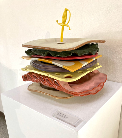 A sandwich made of stoneware showing layers of bread, lettuce, tomato, onion, cheese and ham.