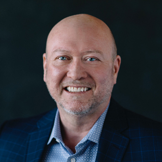 Cohn Drennan is facing forward and smiling. His head is shaven and he wears a light blue shirt under a dark blue jacket.