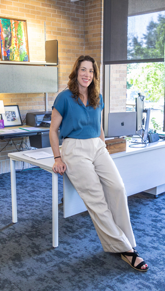 Karen stands in front of a white desk in her office. She has long brown, wavy hair and wears a blue top and beige pants.