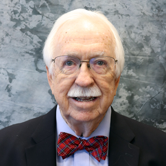 Jack is facing forwar and smiling. He has white hair, a white mustache and wears glasses, a bow tie with a shirt and jacket.