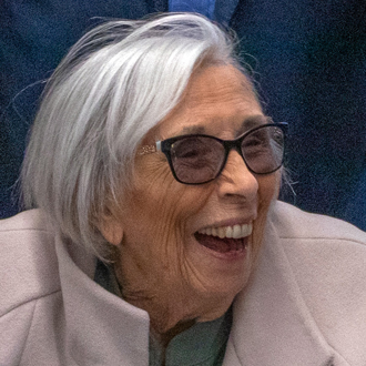 Billie Gough looking to her left and smiling. She has white hair and wears a pink coat.