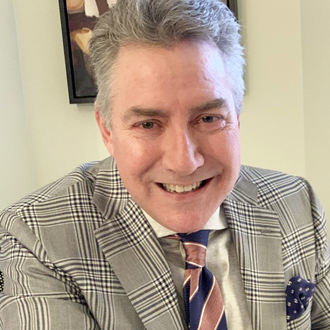 Gordon is facing forward and smiling. He has gray hair and wears a brown plaid jacket, with a matching shirt and tie.