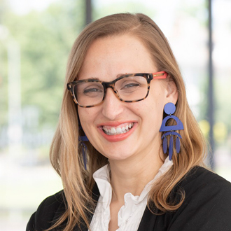 Stefanie is facing forward and smiling. She has long brown hair, wears glasses and a white shirt under a black sweater.