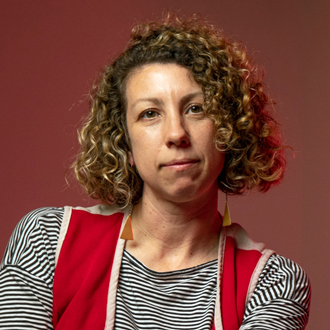 Alicia is facing forward wearing a striped shirt and a red vest. She has brown curly hair.