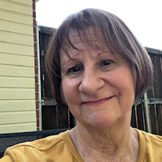 Ellen is facing forward and smiling. Her brown hair is short and she is wearing a gold top.