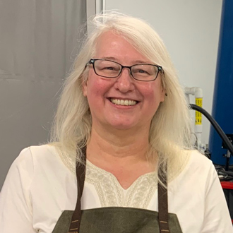 Christa is facing forward and smiling. She has long blonde hair and wears glasses, a beige shirt and brown apron.