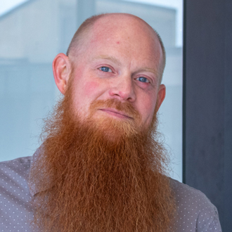 Matt is facing forward and smiling. He has a shaved head, a red mustache, a long beard and wears a gray shirt.