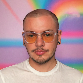 Zak is looking forward and down. His head is saved and he wears a white T-shirt. The background is awash in pastel colors.