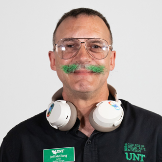 Jeff is facing forward and smiling. He has short brown hair and a green mustache and wears a black button-down shirt.