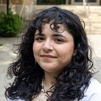 Monda is smiling. She has long, curly, dark hair. The background is an outdoor setting.