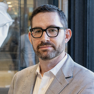 Peter is facing forward. He has dark hair and a beard and mustache and wears a white shirt under a beige jacket.