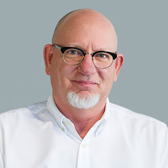 Owens is facing forward and smiling. His head is shaved, he wears glasses, has a white beard and wears a white shirt.