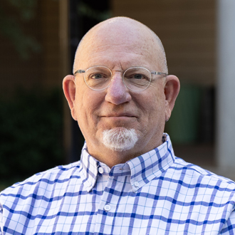 Keith is facing forward and smiling. He is bald, wears black glasses and has a white beard.
