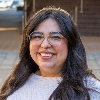 Lyssia is facing forward and smiling. She has long dark hair and wears glasses and a white shirt.