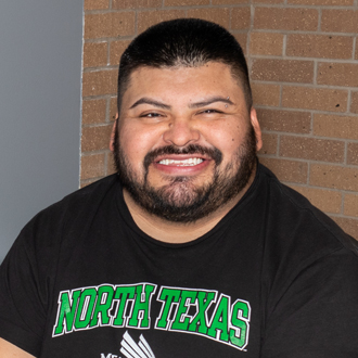 Erasmo is facing forward and smiling. He has short dark hair, a mustache and beard and wears a black North Texas T-shirt.