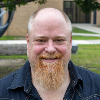 Justin facing forward and smiling. He has a red and gray beard and wears a blue shirt.