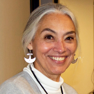 Johnnie is facing forward and smiling. Her gray and white hair is pulled back, she wears silver earrings, a white turtleneck shirt and a beige jacket.