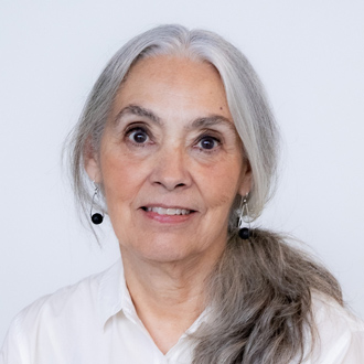 Johnnie is facing forward and smiling. Her gray and white hair is pulled back, she wears silver earrings, a white turtleneck shirt and a beige jacket.