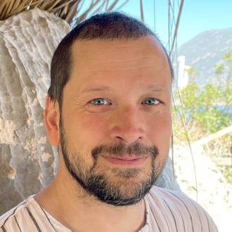 James is facing forward and smiling. He has short dark hair, a mustache and beard and wears a striped shirt.