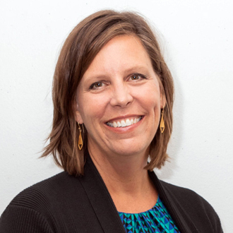 Jeannine facing forward and smiling. She has brown shoulder-length hair and wears a blue shirt under a black jacket.