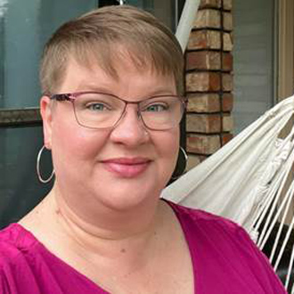 Angela is facing forward and smiling. She has short hair, wears glasses, hoop earrings and a magenta top.