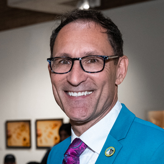 Hepi is facing forward and smiling. He has short brown hair and wears glasses, a white shirt and tie with a turquoise-blue jacket.