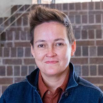 Ursula is facing forward and smiling. She has short brown hair and wears a denim jacket over a rust-colored shirt.