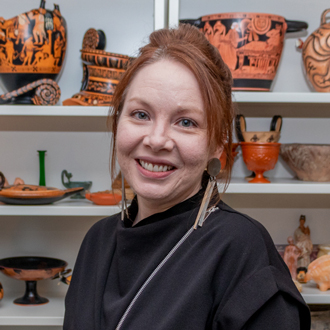 Neville is facing forward and smiling. Her red hair is pulled back and she wears a black top. The background shows reproductions of ancient artifacts.