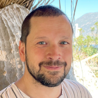 James is facing forward and smiling. He has short dark hair, a mustache and beard and wears a striped shirt.