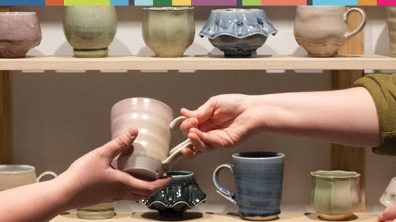 Two shelves of ceramic cups with one hand giving a cup to someone else's hand,