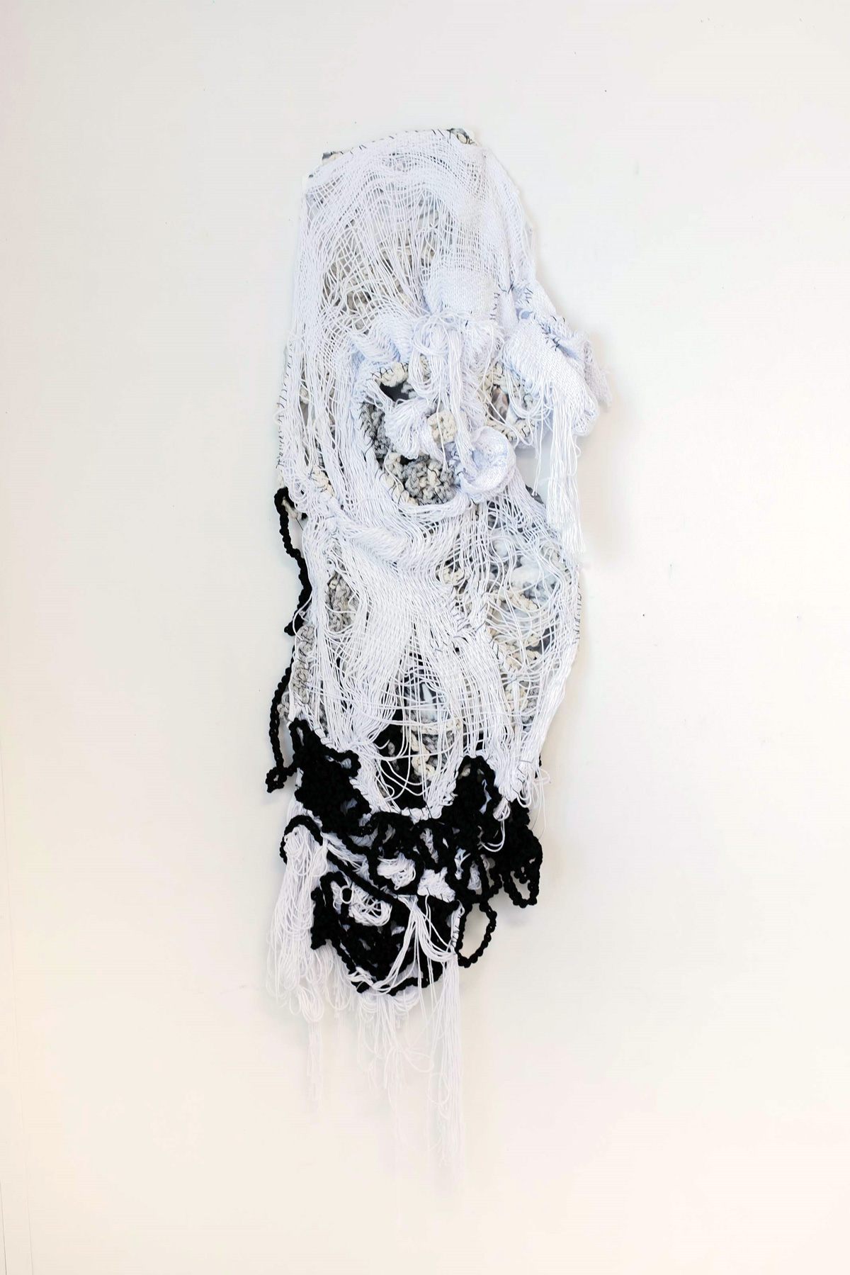 A woven gray, black, and white fiber sculpture hanging vertically on a white wall.