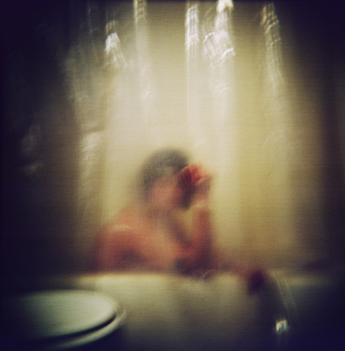 Blurry color photographic image of a figure in a bathtub behind a shower curtain, edge of toilet seen.