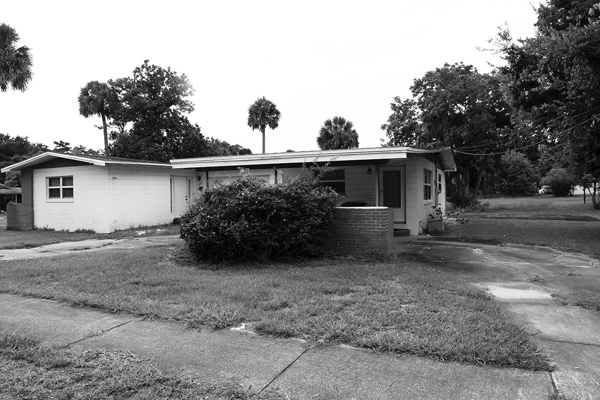 Black-and-white photograph of a one-story home with sidewalks, a carport, and a driveway. There are four Palm trees behind it. 