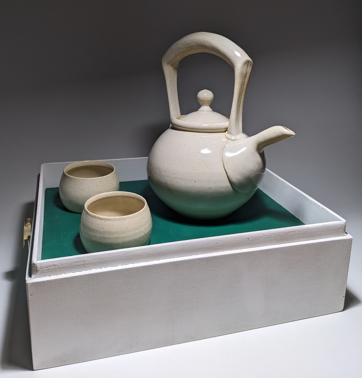 Off-white teapot with matching cup sitting in a white box with a green interior.