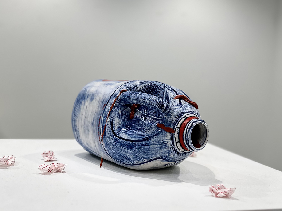 Ceramic sculpture of a blue and white jug lying on its side. Pieces of bubble gum lay on the table around it.