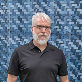 Kenneth 'Kacey' Close III, white hair and beard, glasses, black shirt standing in front of a blue background
