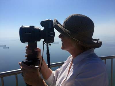 Dornith in profile, wearing a hat, looking through her camera, lake in background with boats