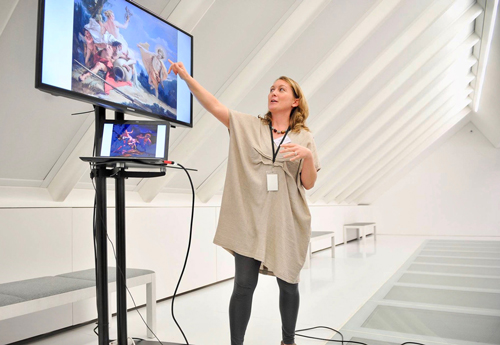 Laura Evans pointing at an oil painting detail that is projected on a large monitor.