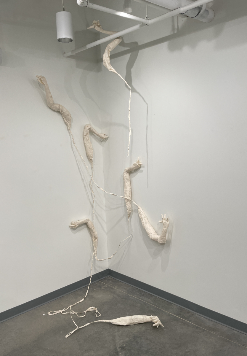 Sculptural arms made of papier mâché that are connected by paper ropes hanging in a white corner area with pipes and lighting at the top.
