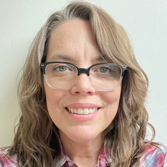 Carla with dark blonde hair and glasses. She is smiling and wearing a red and blue plaid shirt. 