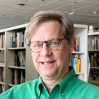 Eric, smiling, wearing glasses, brown hair, blue eyes, book shelves in the background