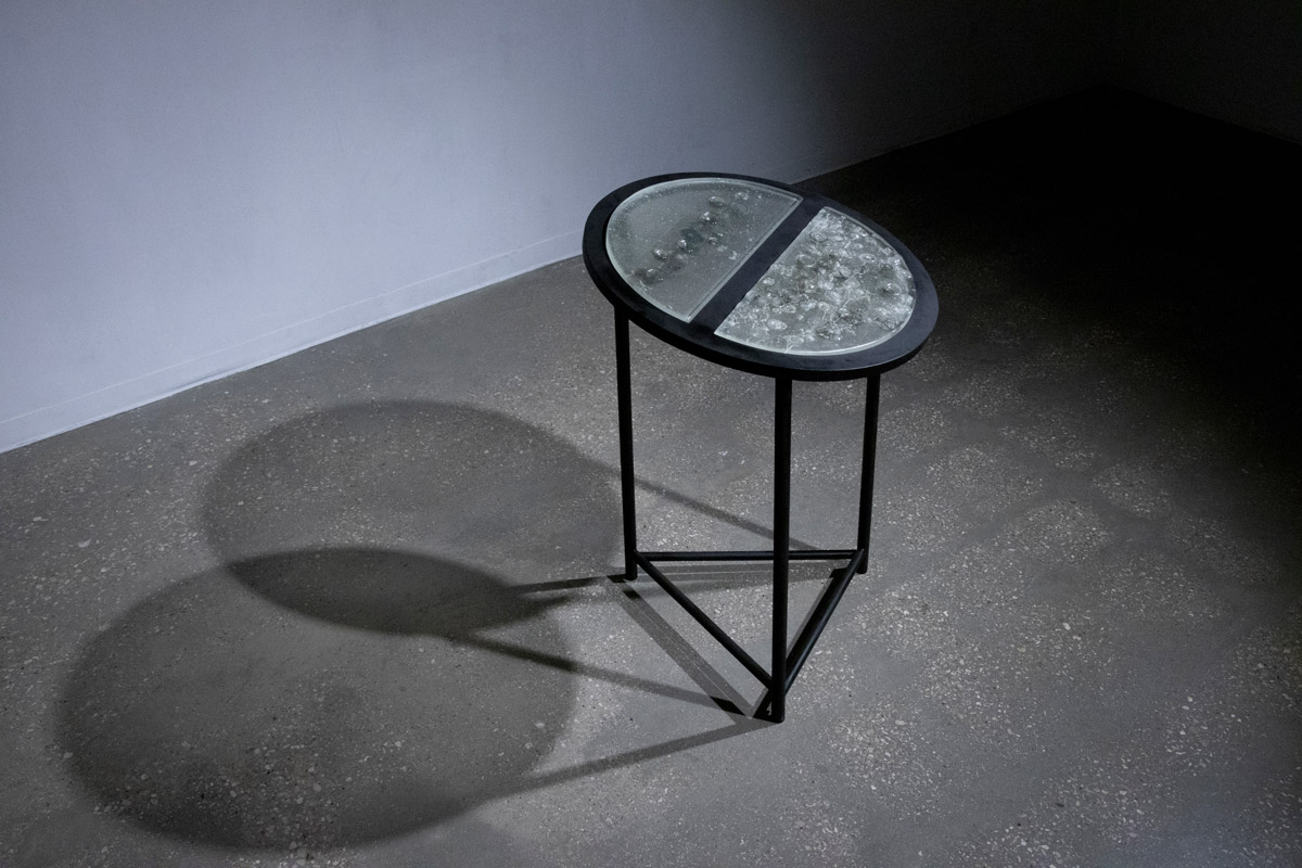 A gallery room with a black circle display table tilted at 35º in the center of it. Two large half-circle glass slabs with plant ash inside them.