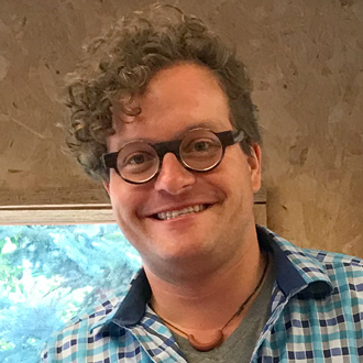 Brooks is facing forward and smiling. He has brown curly hair and wears glasses and a blue plaid shirt