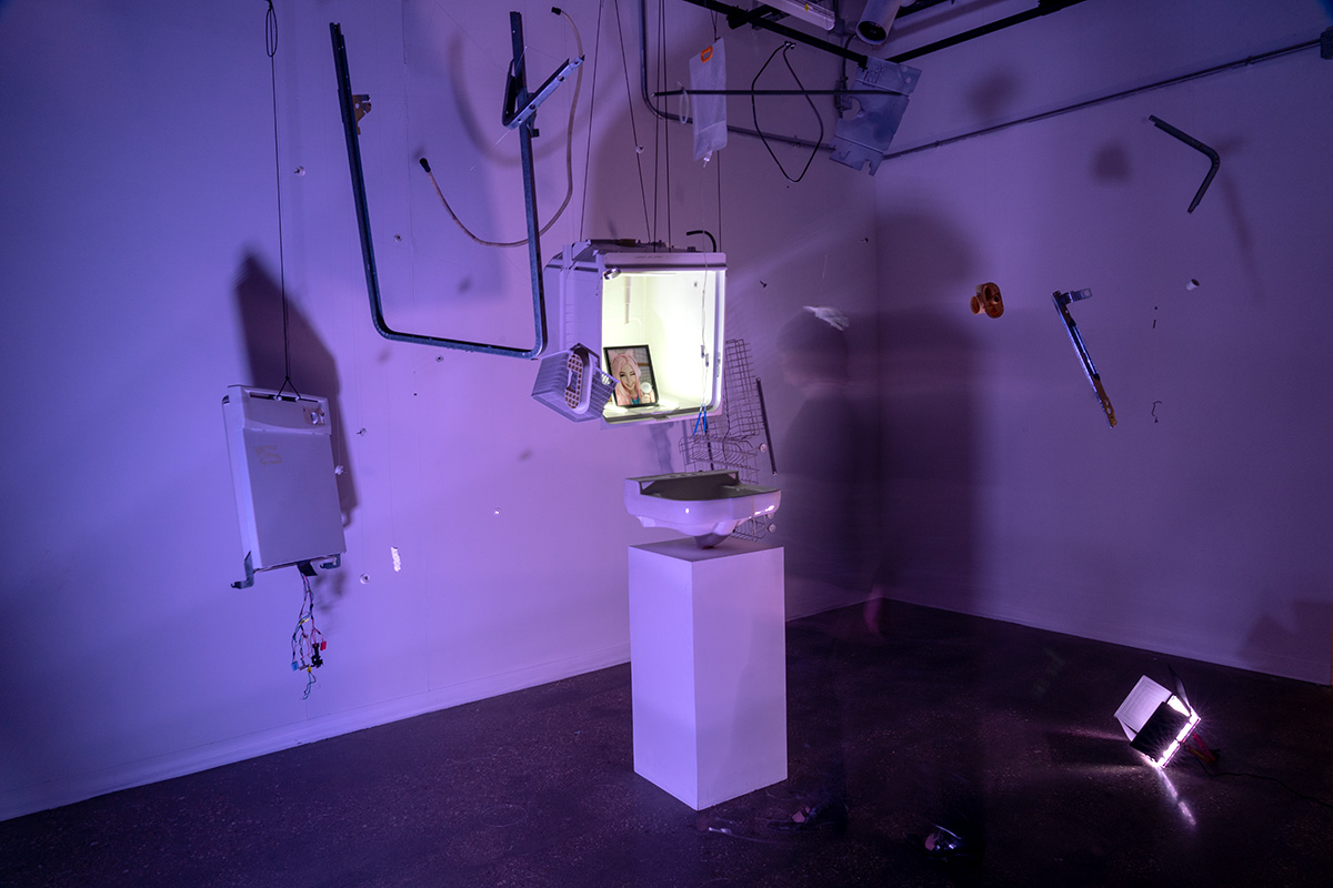 A white room with purple lighting and a dark floor that has various objects hanging from the ceiling and the walls. A box in the center features a photograph of a woman
