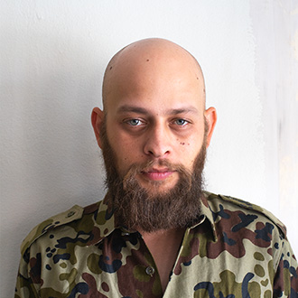 Daniel facing forward, light brown mustache and beard, shaved head, wearing a camouflage shirt. 