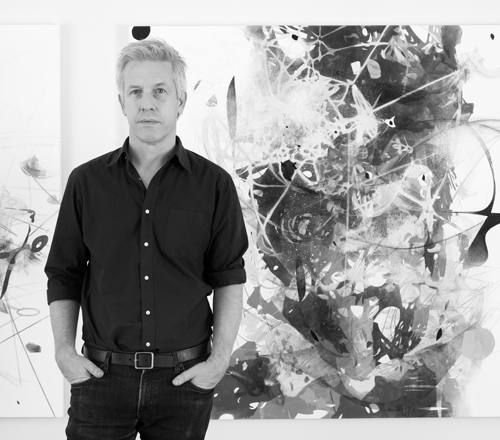 Artist Matthew Ritchie standing in front of an abstract painting, black and white photo