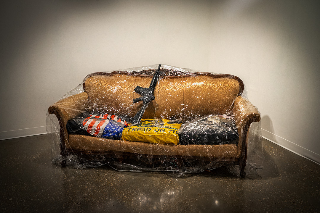 A couch covered in plastic with the cushions covered in U.S. flags and a machine gun leaning on the back.