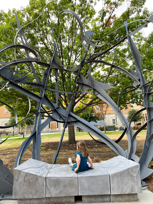A 20-foot tall abstract sculpture with a child on a bench in front of it.